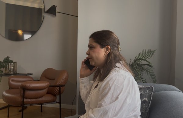 En kvinne med mørkt hår og hvit bluse sitter i en sofa og snakker i mobiltelefon. Rommet er innredet med en brun skinnstol, planter og et stort rundt speil på veggen.