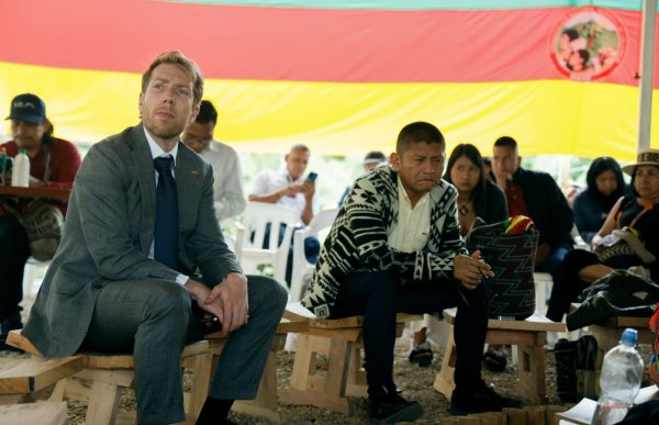 En mann i grå dress, hvit skjorte og blått slips sitter med henda samlet på en tre-krakk. Det sitter flere mennesker der, en ved siden av mannen i dress og flere bak. De hører på et foredrag. 