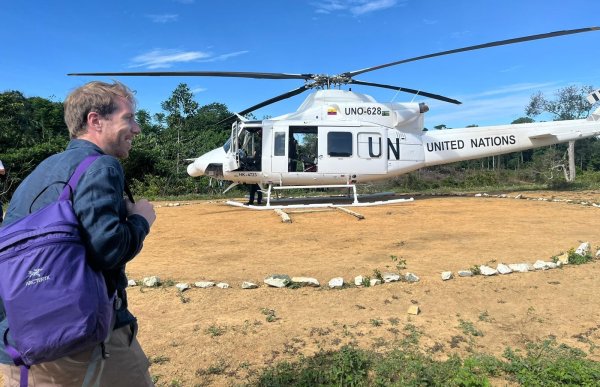 En mann i grønn genser har på seg en lilla sekk.  Han smiler mens han går bortover. Det er blå himmel og sand på bakken, og regnskog i bakgrunn. Det står et hvitt helikopter i bakgrunn. 
