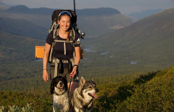 En dame med to hunder går i bånd langs en sti på fjellet. Det er sol og hun har en stor ryggsekk med fiskestang på ryggen.