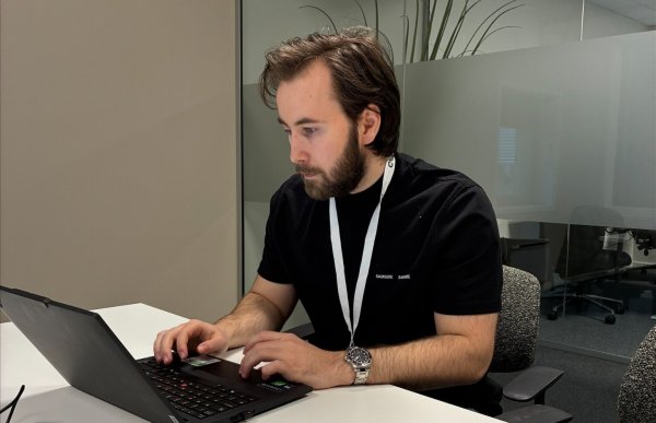 En mann sitter ved et møtebord på et kontor og jobber konsentrert på en bærbar PC. Han har på seg svart t-skjorte, klokke og et ID-kort rundt halsen. Rommet har glassvegger, kontorstoler og et nøytralt, moderne uttrykk. Bildet gir inntrykk av en fagperson i arbeidshverdagen, i rolige og profesjonelle omgivelser.