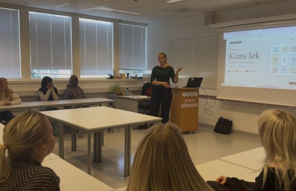 En kvinne med blondt hår i hestehale, mørkegrønn overdel og svart bukse står foran en gruppe elever i et klasserom. Hun gestikulerer mens hun underviser, og på lerretet ved siden av henne vises en digital quiz med tittelen «Kims lek».
