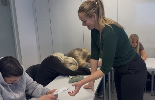 En kvinne med blondt hår i hestehale, mørkegrønn overdel og svart bukse lener seg smilende over en pult i et klasserom. En elev strekker en hånd med en bleie mot henne. Flere elever sitter fremoverbøyd over pultene med lukkede øyne.
