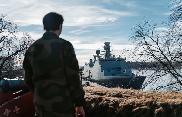 En mann i uniform står i forgrunnen, med ryggen til kamera. Han ser mot et krigsskip som er fortøyd ved kaia. I bakgrunnen er det blå himmel med noen skyer.