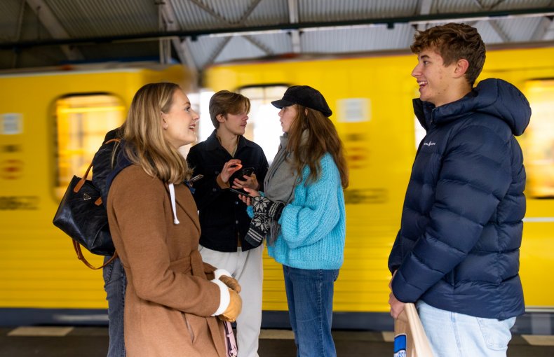 Fem ungdommer står på togstasjonen. En jente har på seg en brun kåpe og en annen har på seg en blå genser. En gutt har på seg en mørkeblå jakke, en annen har på seg en mørk jakke og lyse bukser. En person i mørke klær er lite synlig. Bak ungdommene er det en gul togvogn.