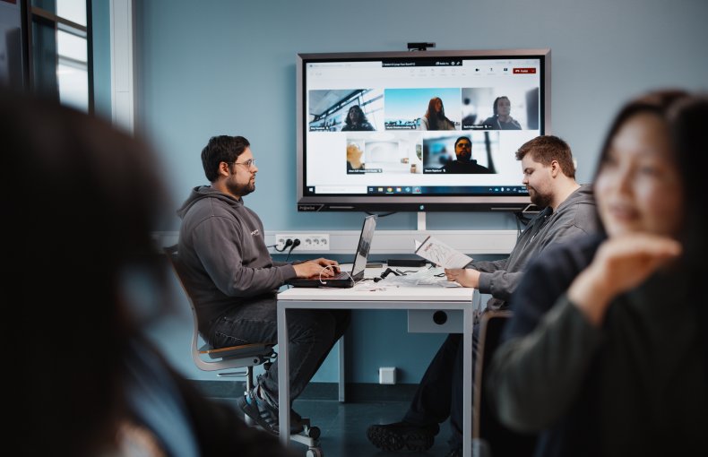 To menn sitter på hver sin side av et bord å jobber med pc, i bakgrunnen er det en skjerm som viser at flere studenter deltar digitalt i undervisningen på zoom
