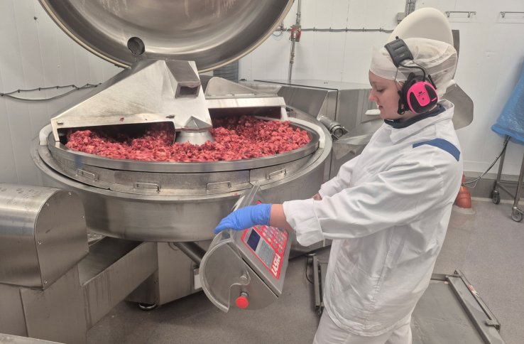 En dame står i hvitt arbeidstøy og har på seg hørselsvern. Hun befinner seg i et matproduksjonslokale.