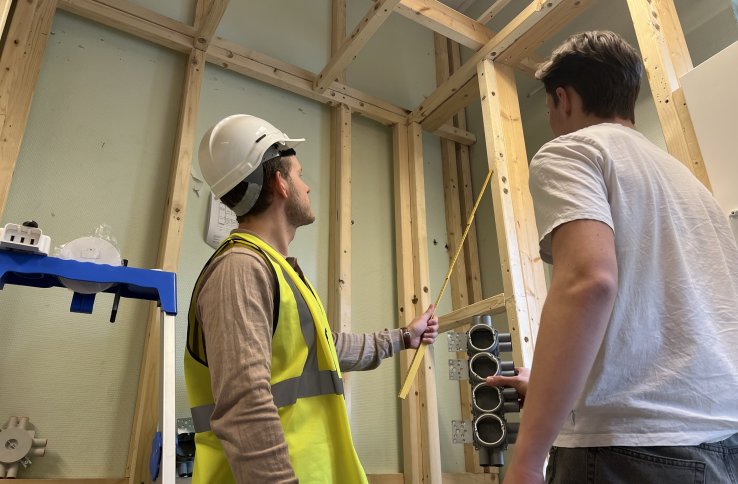 To personer står i et rom hvor reisverket i tre vises. Den ene personen har på seg gul refleksvest og hvit vernehjelm og peker en tommestokk mot den ene planken. Den andre personen har på seg hvit t-skjorte. Bilde er tatt nedenfra. De ser bort fra kamera.