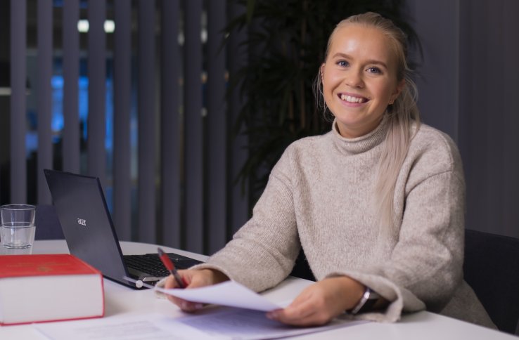 En ung kvinner med lyst hår i hestehaler sitter ved et bord og smiler, hun har et ark i hendene og Norske lover på pulten