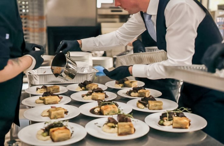 Et kjøkkenmiljø under matservering. Flere tallerkener med forseggjort mat er plassert på et langt bord. En person i kokkeuniform og en servitør i hvit skjorte og vest samarbeider om å legge saus på rettene. Stemningen er profesjonell og travel.