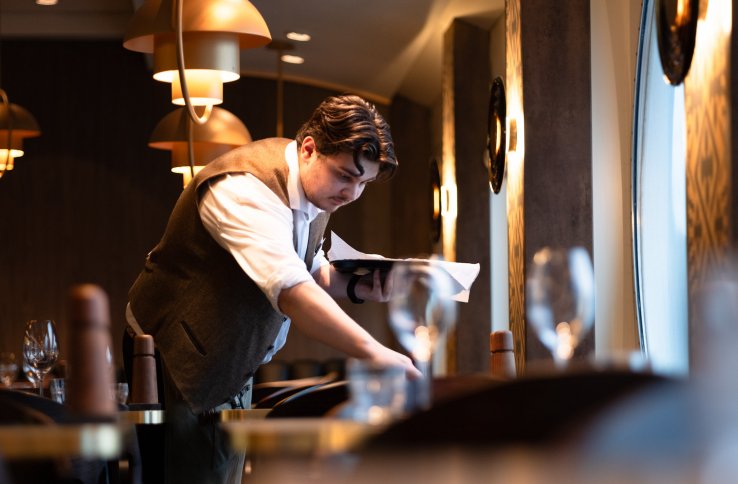 Mann i hvit skjorte og vest dekker et bord. Han er på en restaurant.
