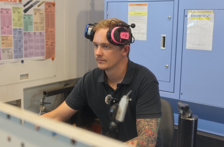 En mann sitter ved en dataskjerm i et industriverksted. Han har et rosa og svart hørselsvern på hodet og bruker en mørk arbeids­pique. I bakgrunnen står store CNC-maskiner og oppslag med tekniske tabeller. Personen jobber konsentrert foran skjermen.