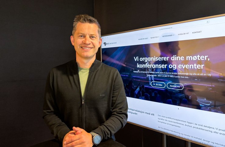 Mann med kort, mørkt hår smiler mot kamera. Han har på seg svart genser med en lys t-skjorte synlig i åpningen. En TV står på i bakgrunnen, inne på kontoret hans.