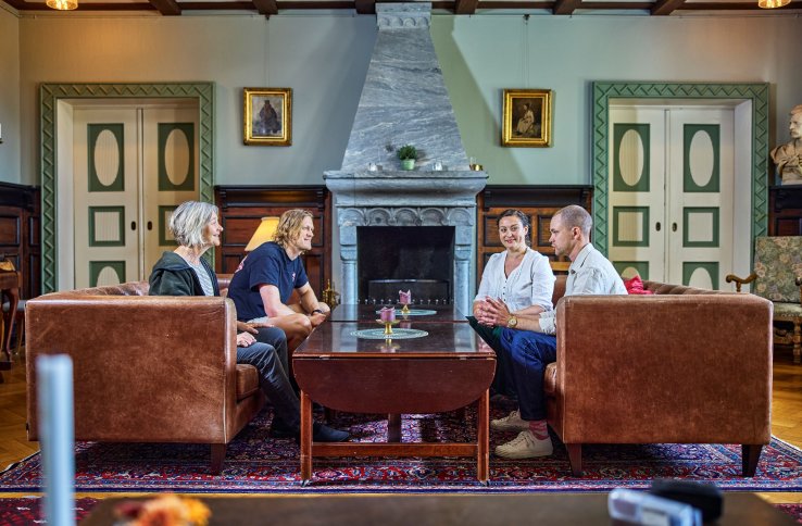 Fire personer sitter i et staselig rom i et hus. Det er en stor peis og fine, grønne dører på hver side. Det er to damer og to menn, de sitter to og to i brune skinnsofaer og prater. 