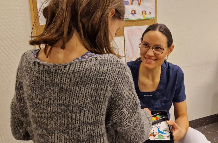 En dame med mørkt hår og briller, ikledd blå t-skjorte smiler til en ung jente med grå genser og brunt hår. Det er tegninger på veggen i bakgrunnen.