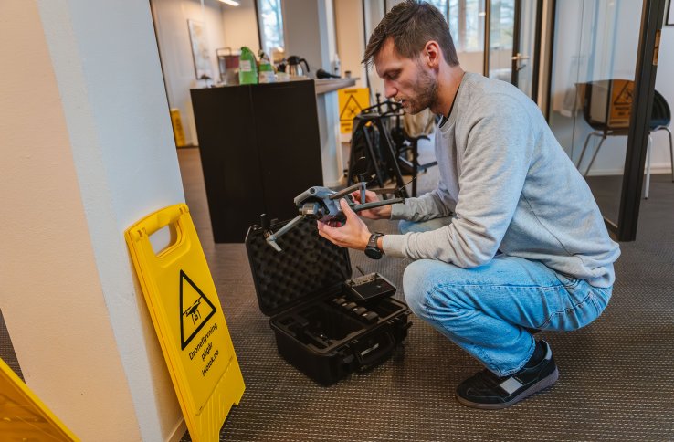 En mann med skjegg, iført olabukse og en lysegrå genser, sitter på huk og setter sammen en liten drone. Ved siden står et gult skilt med teksten "droneflygng pågår".
