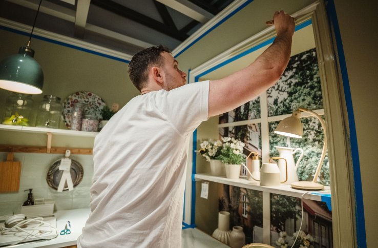 En maler står på et kjøkken og strekker seg til toppen av en list. Listen ligger rundt et vindu, og er maskert med blå teip.