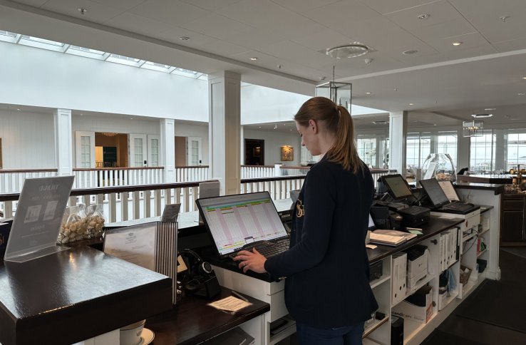 En kvinne står bak en lang skranke. Hun har brun hestehale og har på seg en mørkeblå dressjakke. Hun står foran en PC som viser en slags tabell. Foran henne kan man se et hvitt rekkverk. Ved siden av henne er flere andre skjermer og en hylle med diverse permer og annet utstyr.