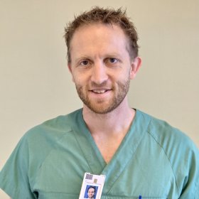 En mann med brunt hår og skjegg smiler. Han har på seg en grønn sykehusskjorte som leger har på seg. På skjorten henger avgangskortet hans, og på høyre side er det en lomme på skjorten hvor det er en penn som stikker opp. Bak mannen er det en beige vegg.