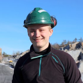 Ung, smilende mann med grønn hjelm og mørk genser. I bakgrunnen skimtes stein, trær og en blå himmel. 