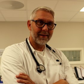 Mann med grått hår og briller har et stetoskop rundt halsen og er kledd i hvitt. 