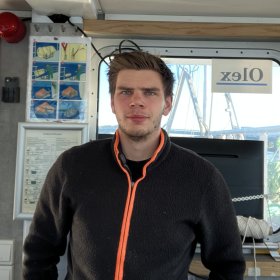 En mann med svart jakke står i en båt, han har mørkt hår og ser rett inn i kamera