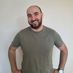 Portrettfoto av skoleassistent Tommy Egeland Mann med mørkt, kort skjegg og en grønn t-skjorte står foran en lys vegg og smiler til kamera.