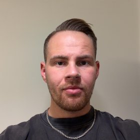 En mørkhåret mann med brune øyne og piercing i nesen ser inn i kamera