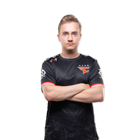 En ung mann står med armene i kors mot en hvit bakgrunn. Han har kort, lyst hår og et nøytralt ansiktsuttrykk. Han har på seg en svart e-sportdrakt med røde detaljer og en tydelig laglogo på brystet.