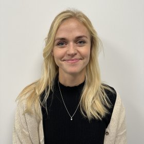 Lyshåret dame med svart t-skjorte og hvit jakke over smiler til kamera med en hvit vegg i bakgrunnen