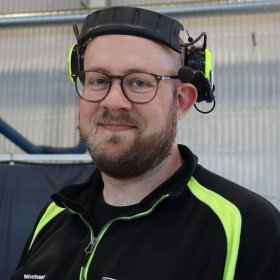 Ung mann med helskjegg og briller smiler til fotografen. På hodet har han hørselvern og han er ikledd en svart genser med gule striper på skuldrene. 