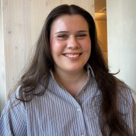 En jente med brunt hår og stripete blå skjorte som smiler bredt.