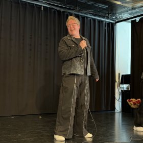 Mann med kort, blondt hår, iført svart bukse og svart jeansjakke, står på en scene og synger mens han holder en mikrofon. Bakgrunnen er mørk.
