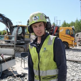 En ung mann iført gul refleksvest, vernehjelm og hørselsvern står på en byggeplass og smiler mot kamera. I bakgrunnen står det en gravemaskin og en lastebil. 