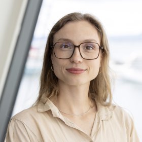 En dame med beige bluse og brunt hår smiler med munnen lukket. Hun har på seg briller. Bak henne er det en lys bakgrunn.