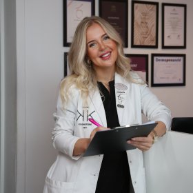 Kvinne med langt blondt hår iført hvit medisinsk frakk holder en svart notatblokk og en penn. På veggen bak henne henger flere innrammede diplomer.