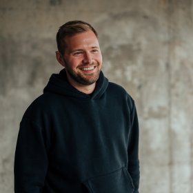 En mann med blondt hår og skjegg og svart hettejakke står foran en grå vegg