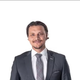 En mann med mørkt, bakovergredd hår og kort skjegg står foran en lys grå bakgrunn. Han har på seg mørk dress, hvit skjorte og slips, og ser rett inn i kamera med et rolig smil.