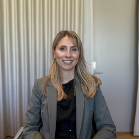 En kvinne sitter vendt mot kamera i et kontormiljø og smiler vennlig. Hun har langt, lyst hår som faller over skuldrene og er kledd i en mørk topp under en grå dressjakke. Bak henne henger lyse gardiner som dekker en vegg eller et vindu, og en dør er synlig i bakgrunnen.