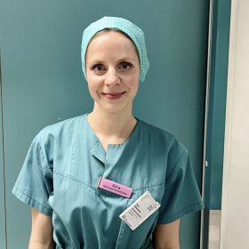Portrettbilde av en dame i grønn operasjonsklær. Hun står foran en grønn dør. I arbeidsskjorten har hun et skilt som sier hun er operasjonssykepleier 