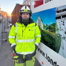 En anleggsrørlegger står ute på en byggeplass, kledd i gul refleksjakke og hjelm med hodelykt. Han ser rett mot kamera og står ved siden av en arbeidsbil med firmalogo og bilder av ferdige prosjekter.