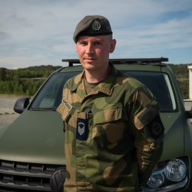 En person i militæruniform med beret står foran et militært kjøretøy og ser mot kameraet. Bakgrunnen viser åpent landskap og en parkeringsplass.