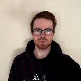 En mann med brunt hår og briller og svart hettejakke står foran en lys vegg. 