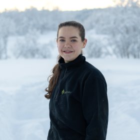 Ei dame i mørkeblå genser. Hun smiler til kameraet. Hun er ute. Det er snø bak henne.
