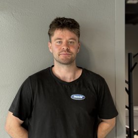 En mann står foran en grå vegg og ser mot kameraet. Han har på seg en svart T-skjorte med Hasle-logo på brystet.