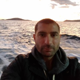 En mann med mørkt kort hår tar en selfie på en båt i solnedgangen. Bak ham er sjøen og to lave øyer i horisonten, mens himmelen er lys med noen mørke skyer.