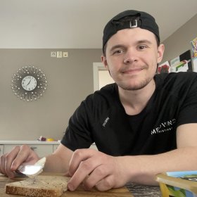 En ung mann i svart t-skjorte og svart caps sitter ved en kjøkkenbenk og smører pålegg på en brødskive