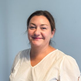 Kvinne i hvit bluse står innendørs og smiler til kamera foran en gråblå bakgrunn