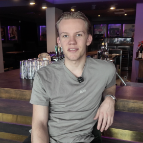 En blond mann med grå t-skjorte sitter lett henslengt foran en bardisk. Bak baren står det diverse flasker og glass