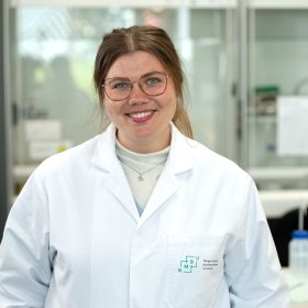 En kvinne i briller og hvit arbeidsfrakk. Hun står i et laboratorium, med arbeidsutstyr og skap i bakgrunnen.